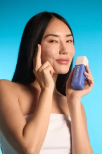 Mulher asiática segurando o Anti-Aging Peptide Cream e passando no rosto, em um fundo azul. 
