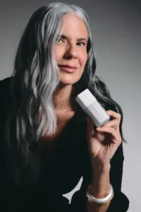 Mulher branca, usando roupa preta, em um fundo claro, segurando o Eye Cream próximo ao rosto.