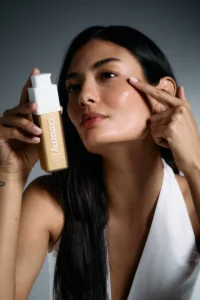 Mulher asiática, usando roupa branca, em um fundo claro, segurando a Vitamina C Gold e passando no rosto.