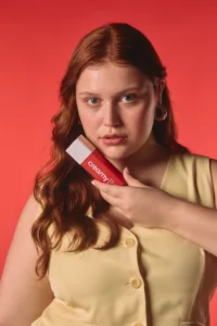 Mulher branca e ruiva, com roupa amarela, em um fundo vermelho, segurando o Sérum Facial Retinal próximo ao rosto.
