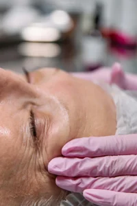 Mulher deitada, usando touca no cabelo, com olhos fechados. Nos olhos, rugas e marcas de expressão, com mãos com luvas apalpando o rosto. 
