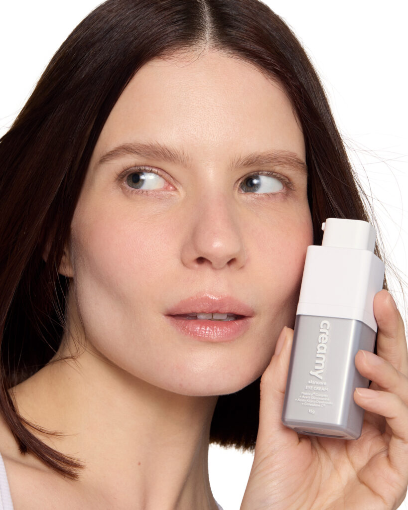 Cuidados com área dos olhos: como montar uma rotina para a região mais delicada do rosto 2 Mulher branca, com olhos claros e cabelo escuro, segurando Eye Cream da Creamy perto do rosto.