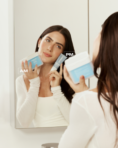 Poros dilatados: por que acontecem e como tratar? 5 Jade Picon com Pore Refiner e Ceramide Skin Repair ao lado do rosto, enquanto se olha no espelho